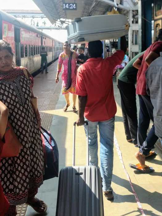 Indien Zugreisen: alles Wissenswerte von der Buchung über Zugklassen bis zur Reise MANU SARONA • health mind spirit • Dein Blog für ganzheitliches Wohlbefinden Ein indischer Gepäckträger mit einem Koffer auf dem Kopf und einen kleinen Trolley ziehend auf einem Bahnsteig mir einigen Personen neben einem stehenden Zug